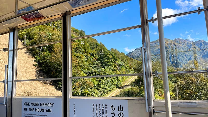 ロープウェーの窓越しに見える山の斜面と青空、奥に連なる八海山の稜線